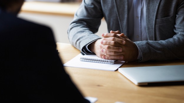 Man with clasped hands at a table during job interview with papers and interviewer in foreground