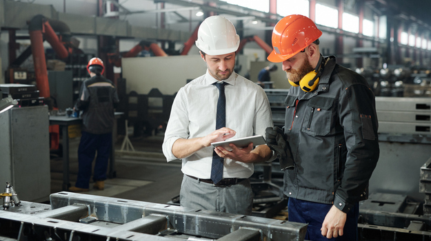 Young master in hardhat and bearded engineer discussing technical sketch on display of tablet in factory workshop